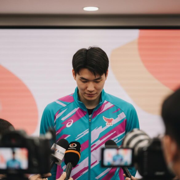 Jugador asiático de voleibol inclina la cabeza en una rueda de prensa, con micrófonos al frente.