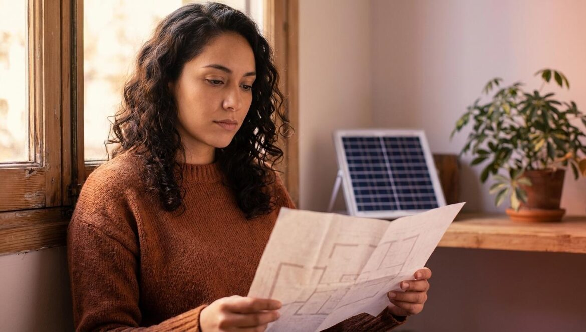 Vida off-grid y crianza: el detalle que lo cambia todo cuando aparece el Estado Mujer joven mira unos planos junto a una ventana rústica con un panel solar al fondo.