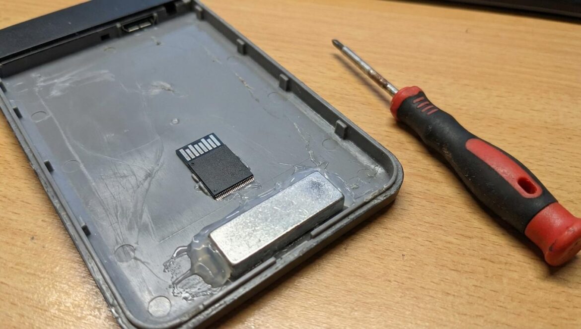 Explota la estafa de discos duros baratos: el truco del peso falso y la memoria que “se inventa” Carcasa de disco duro externo abierta mostrando un chip pequeño y un peso metálico pegado con restos de pegamento.