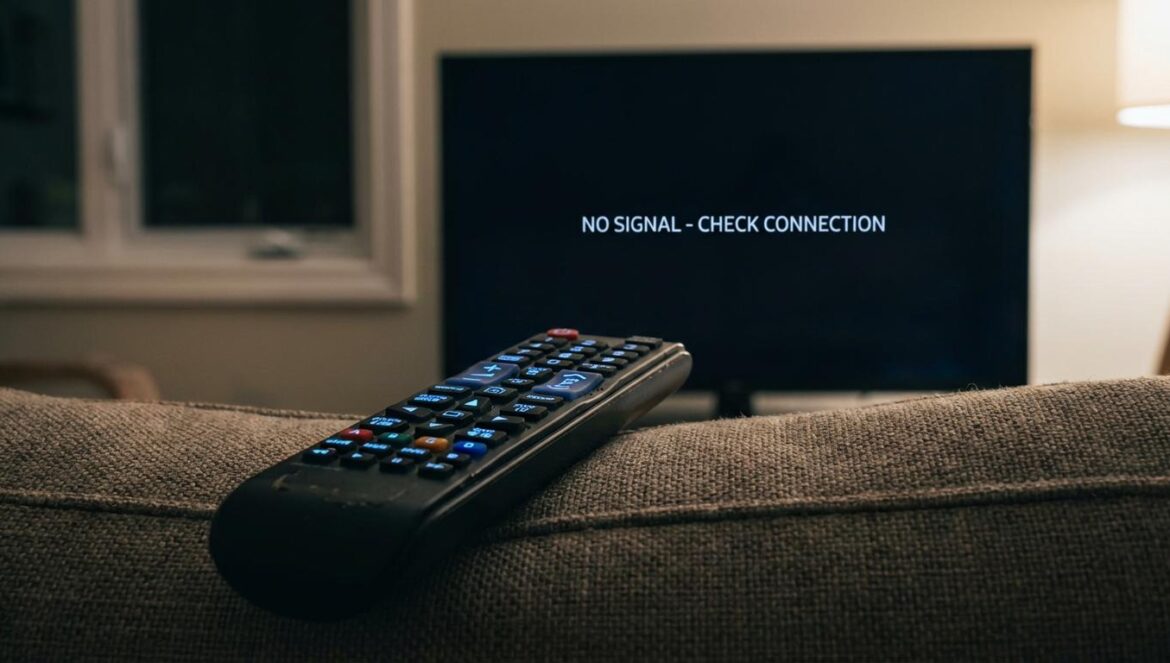 La nueva numeración de la TNT en Francia: el minuto de negro que reconfigura el mando Control remoto sobre un sofá, apuntando a una TV con pantalla negra y mensaje de sistema en una sala con luz nocturna tenue.