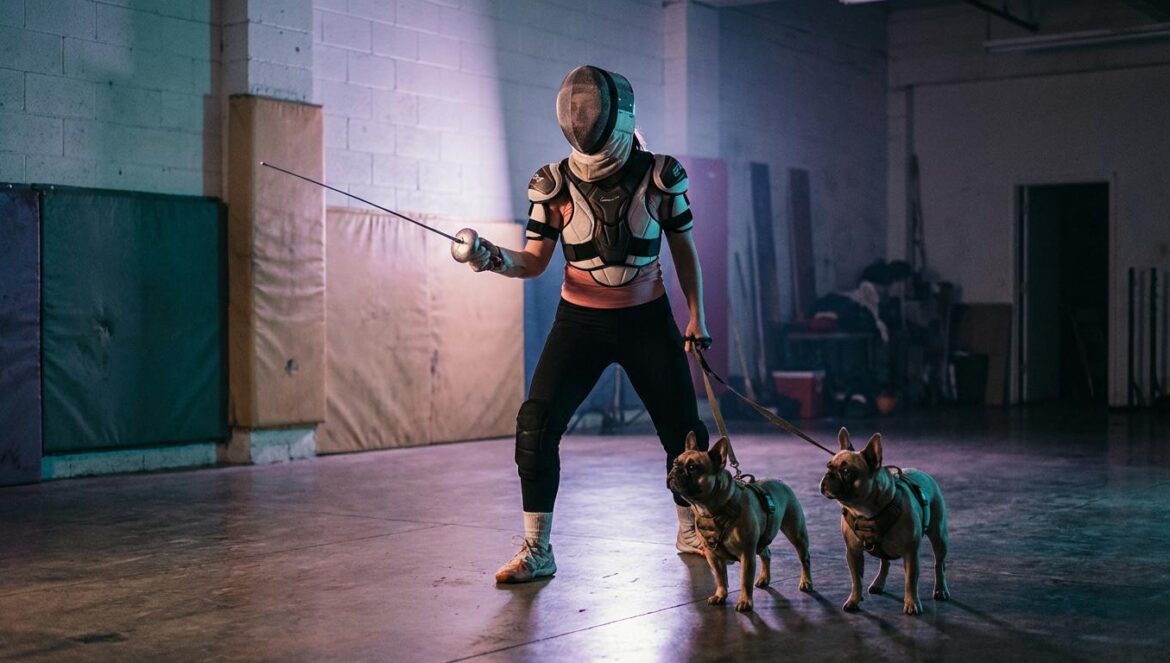 Una artista anónima con máscara de esgrima y protecciones de hockey posa en una sala de entrenamiento, con dos perros atentos a su lado y luz fría cinematográfica