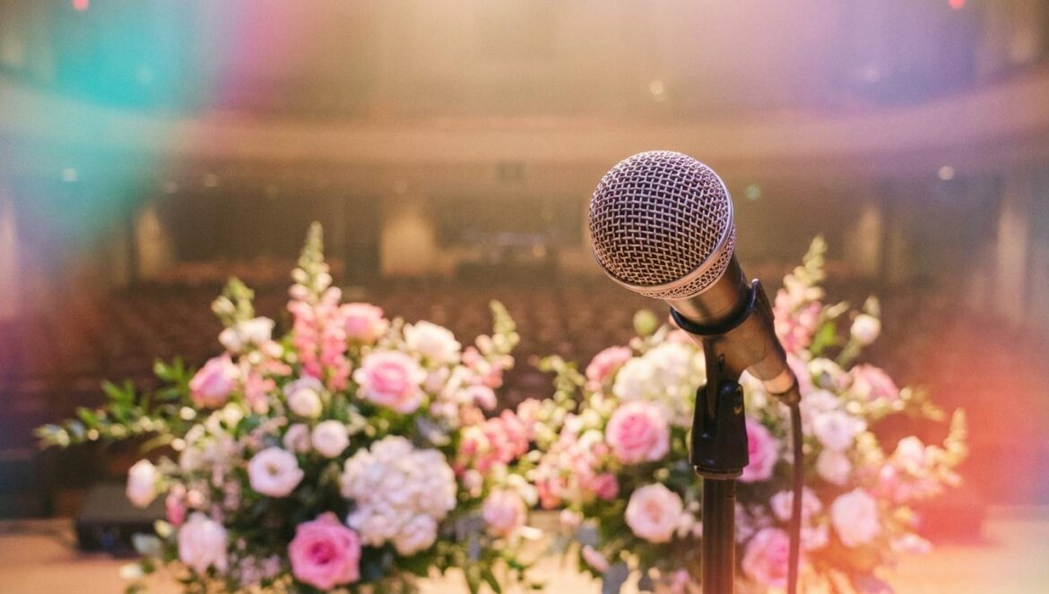 Nam-gung Jin: el poder de la piedad filial en el Trot Micrófono profesional bajo luz cálida con arreglos florales difuminados al fondo, representando una actuación televisiva de música trot.