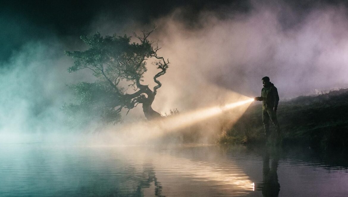 Whispering Water y el giro oscuro de Kim Hye Yoon en el cine Vista nocturna de un embalse brumoso con un árbol retorcido iluminado por una linterna, capturando una atmósfera de terror cinematográfico.
