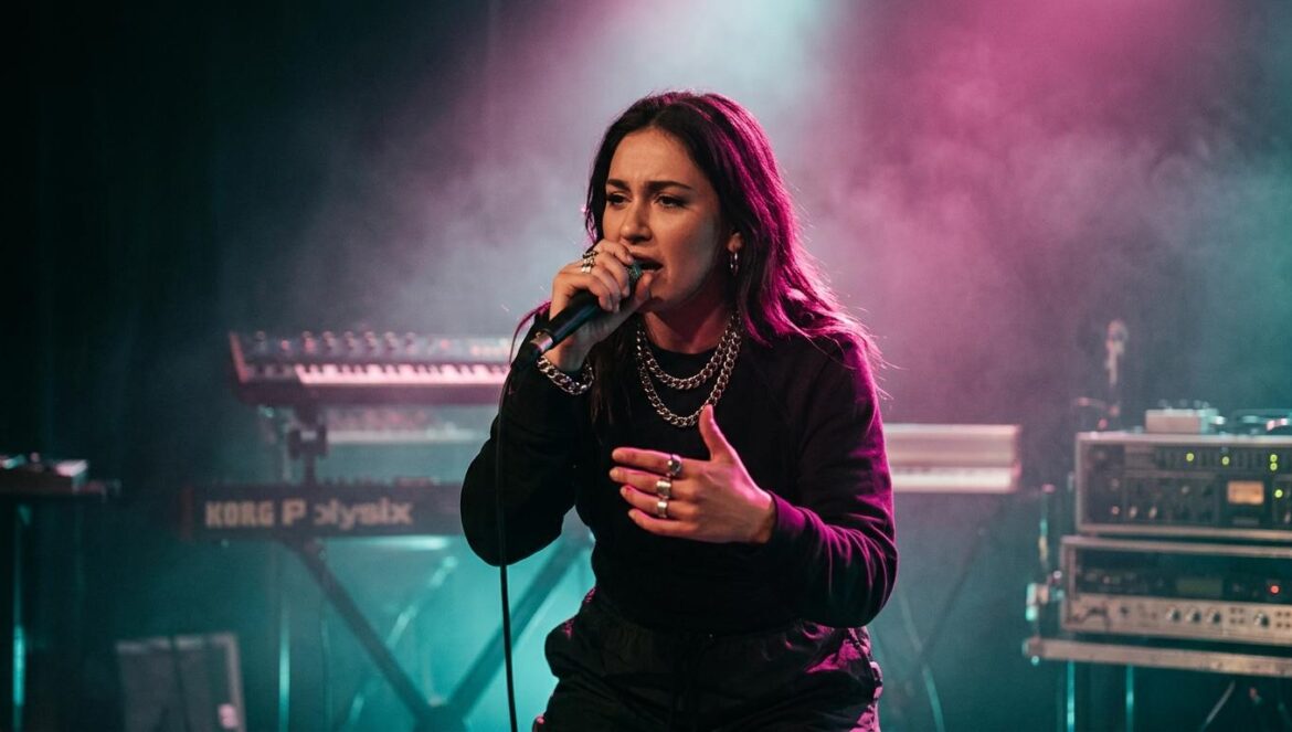 Angel Money y el cansancio de vender hedonismo en internet Una rapera actúa en un escenario de club oscuro y lleno de humo, iluminada por luces de fondo magenta y sombras turquesa con cadenas metálicas.