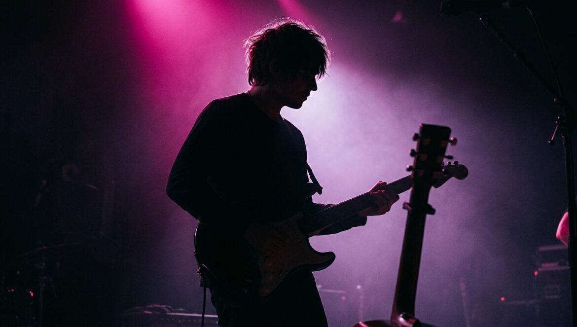 Silueta de un músico sobre el escenario con iluminación magenta de fondo y neblina lila, destacando el mástil de una guitarra acústica en primer plano.