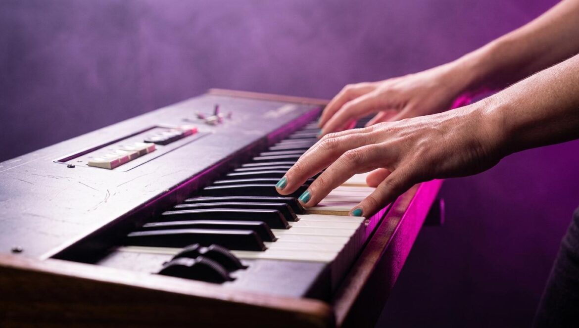 Primer plano de un sintetizador analógico clásico iluminado por luces de escenario magenta y violeta, con manos tocando las teclas en un ambiente retro.
