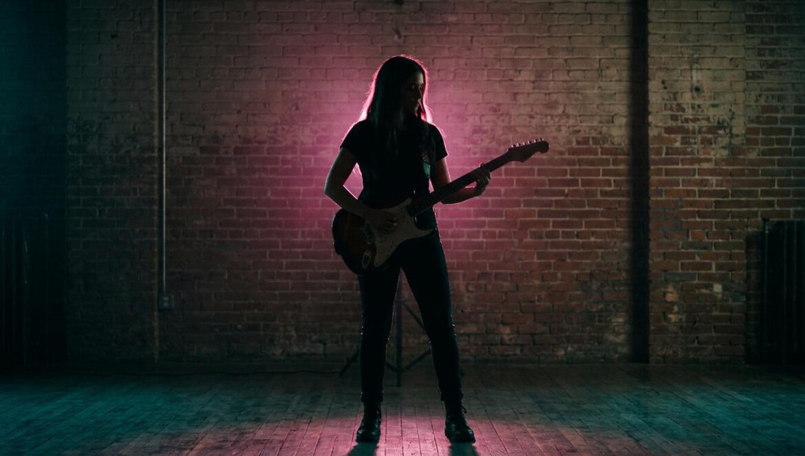 Silueta de un músico sobre un escenario iluminado con tonos turquesa y magenta, con fondo de ladrillo industrial.